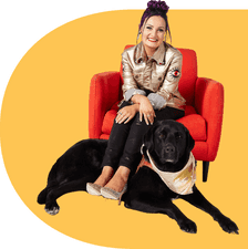 A brown haired young woman in a gold jacket, black pants, and glitter heels smiling at the camera while sitting on a red armchair. Under her legs sits a black dog with a matching gold handkerchief, dwarfing her and the chair.